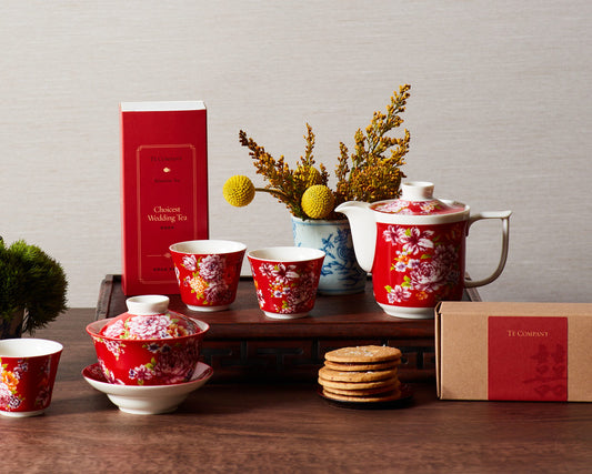 A red floral tea set with cups, teapot, a box of wedding tea, cookies, and flowers.
