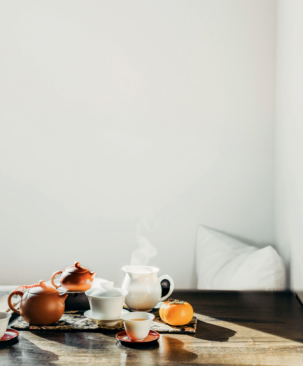 Traditional tea ceremony setup with clay teapots, white porcelain cups, and a persimmon fruit on a dark wooden surface in natural sunlight.