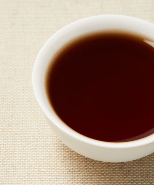 A close-up of blackish-brown tea in a white teacup placed on a beige textured background.