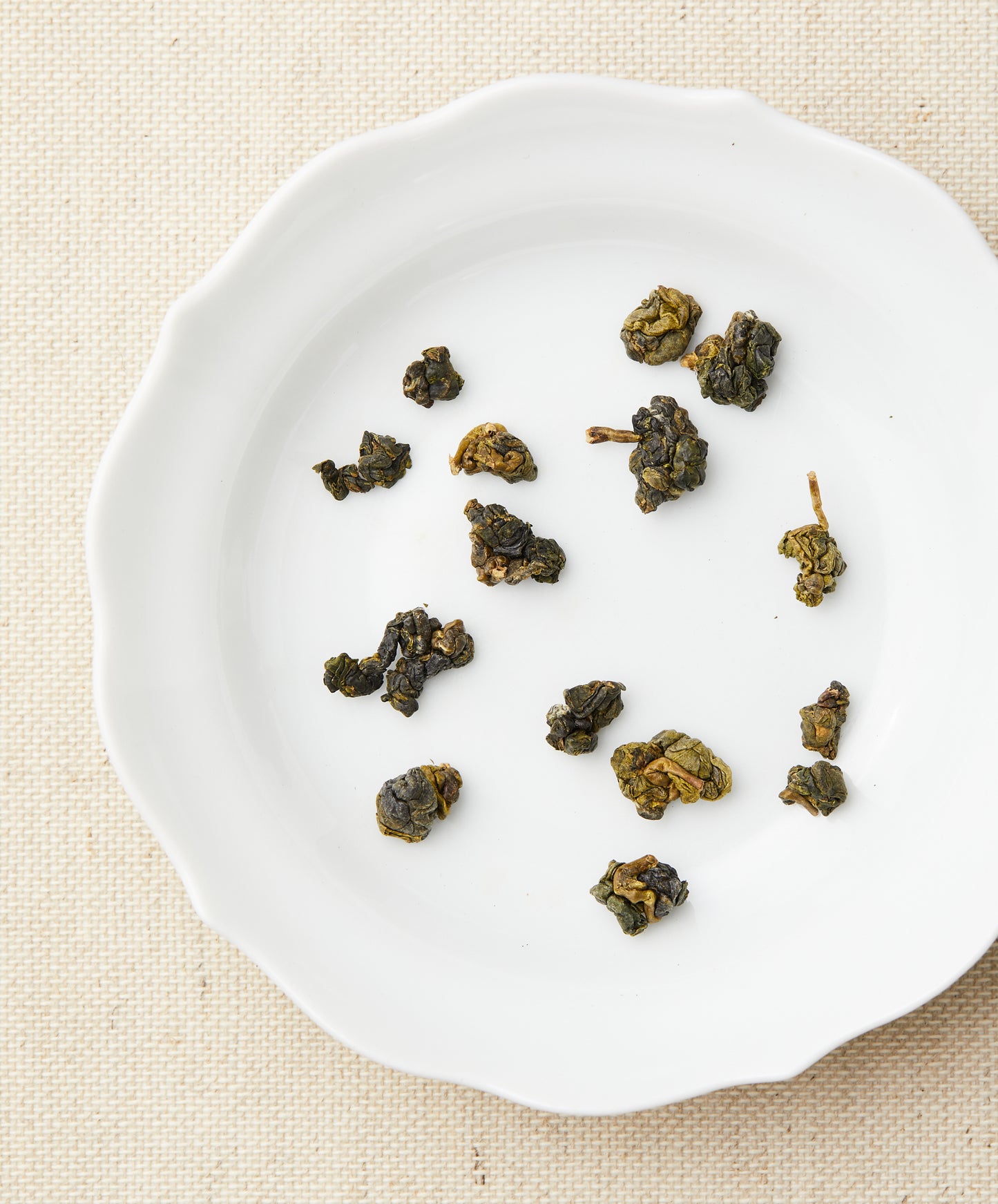 Loose-leaf tea pieces arranged on a white plate, showing tightly rolled green tea leaves against a beige fabric background.