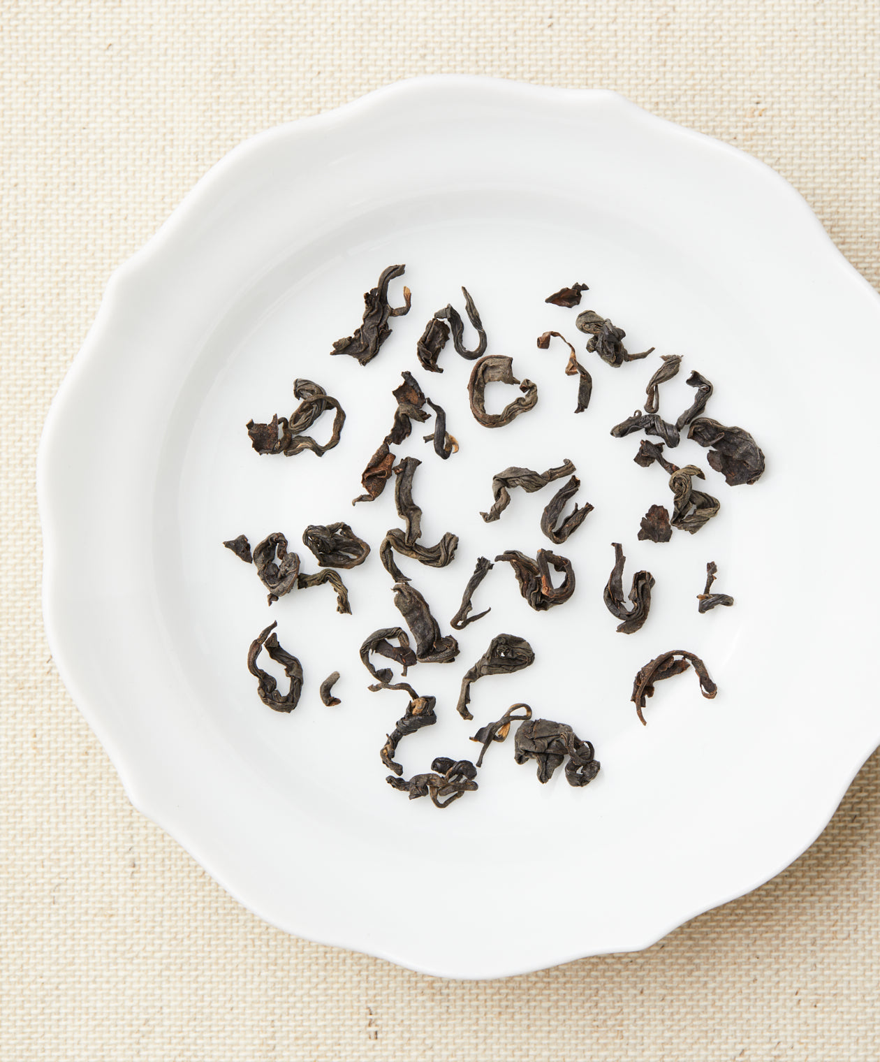 Loose-leaf tea in curled, dark pieces arranged on a white plate against a beige fabric background.