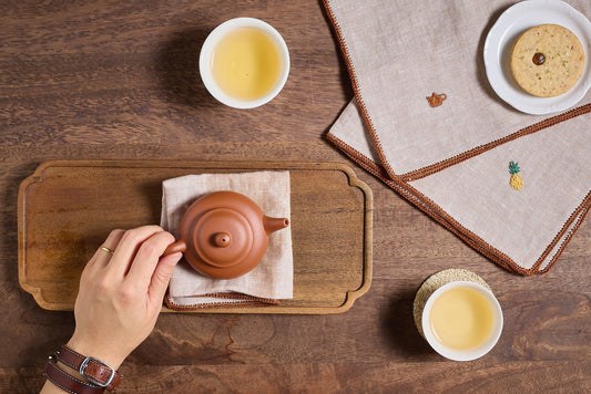 Hand pouring tea with cups and cookie on table.