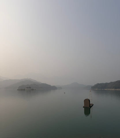 A misty lake scene with distant hills or islands, calm water, and a small floating platform with a vertical structure in the lower right.