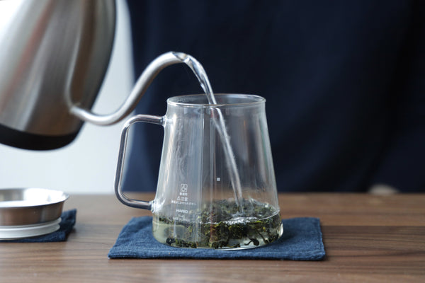 Hot water being poured into a tea brewing pitcher containing loose tea leaves.
