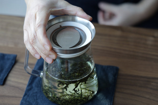 A person placing a metal lid on a tea brewing pitcher