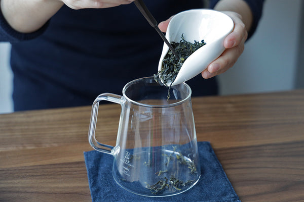 A person adding loose tea leaves to a glass tea brewing pitcher.