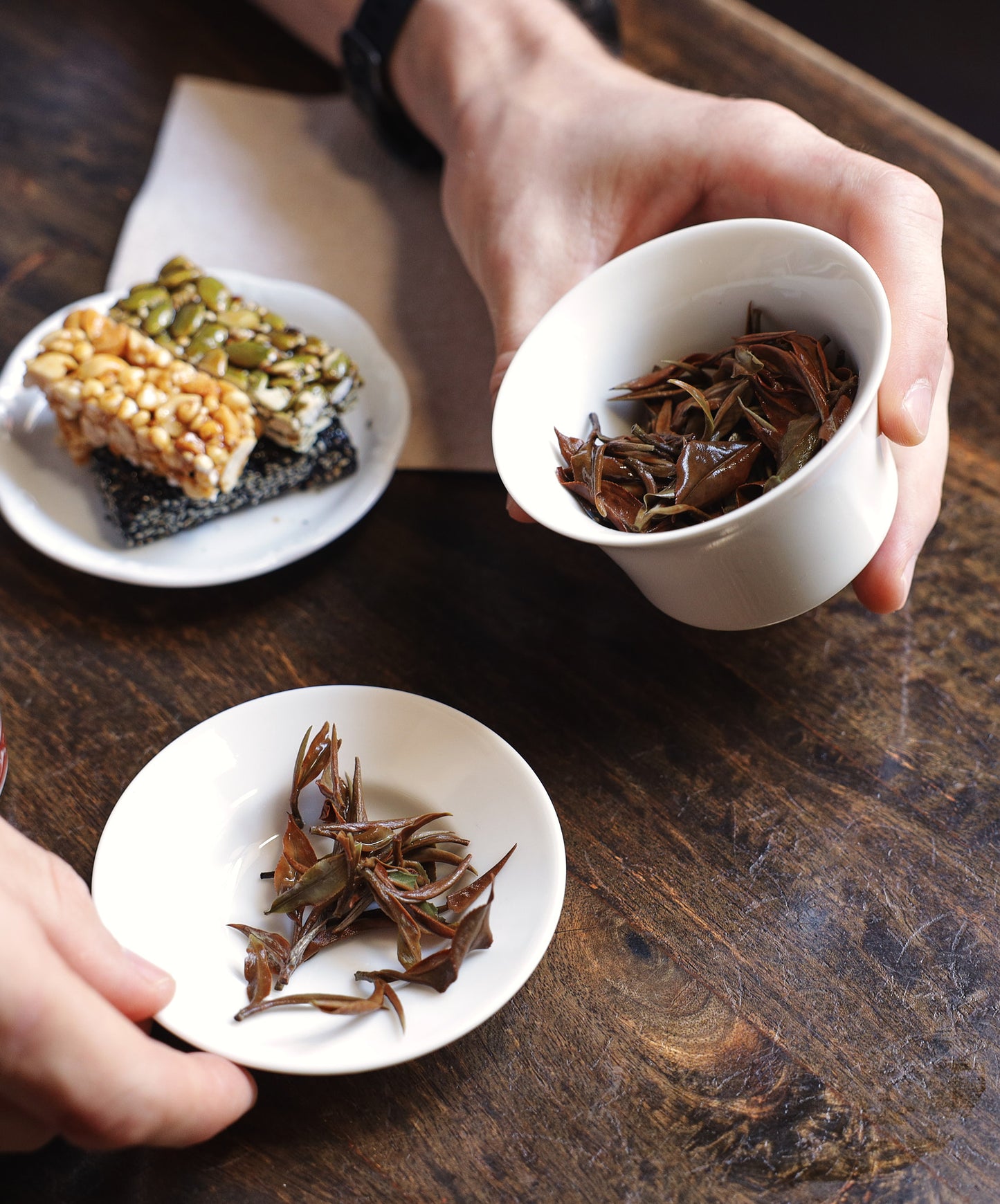Overhead view of steeped tea leaves in white ceramic cups with a sesame and nut bar on a rustic wooden table.