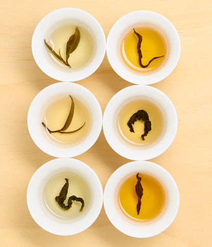 Six small white cups arranged in a 2x3 grid on a light wood surface, each filled with tea and showing different curled tea leaf shapes and water colors.