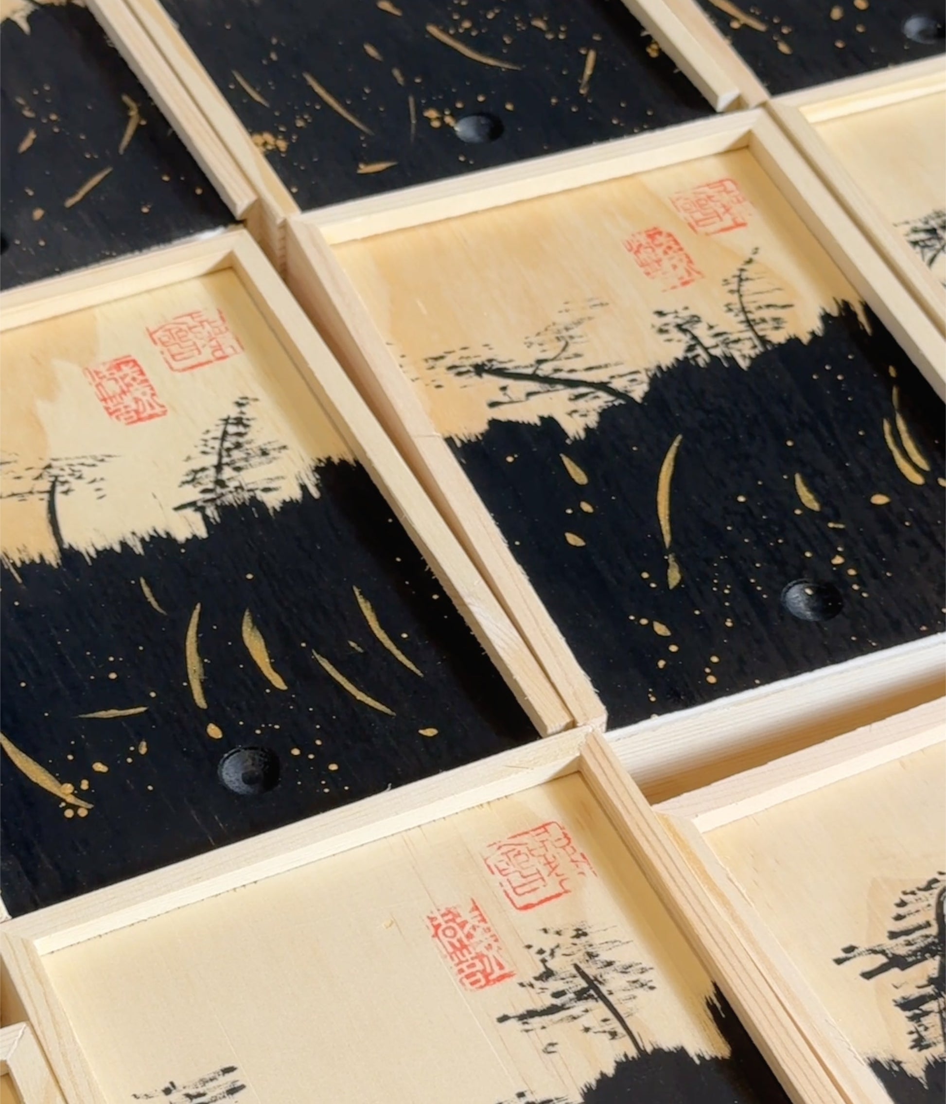 A close-up of multiple rectangular wooden boxes arranged diagonally, each featuring a black and beige landscape-style print with red seal marks and small dark circular details.