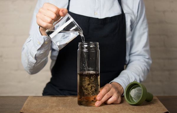 Person pouring water into a glass bottle with tea leaves for brewing.