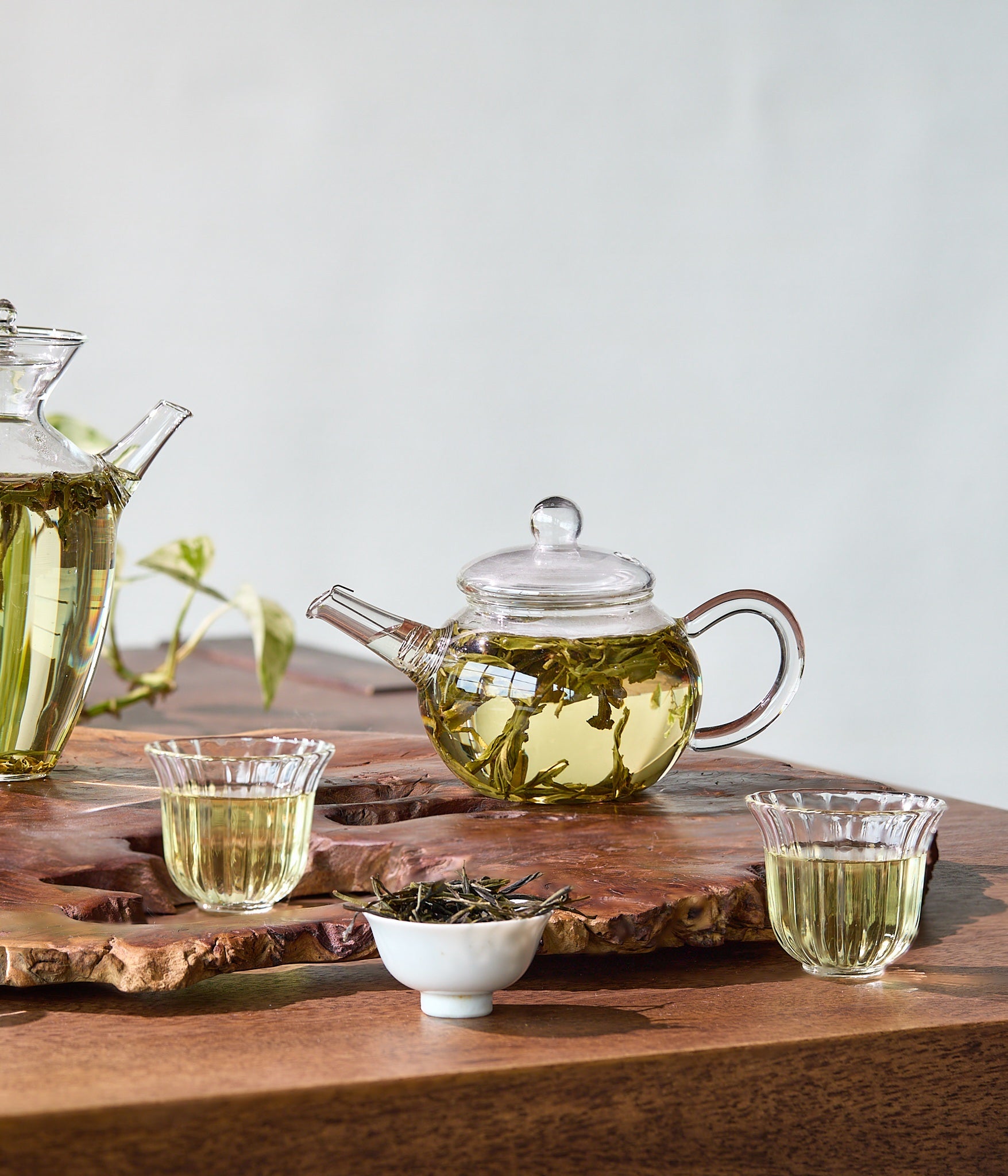 Tea-making setup with glass teapot, cups, and tea leaves on a wooden board.