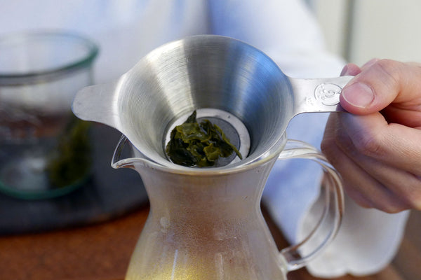 Tea leaves stuck in a metal strainer over a glass pitcher.