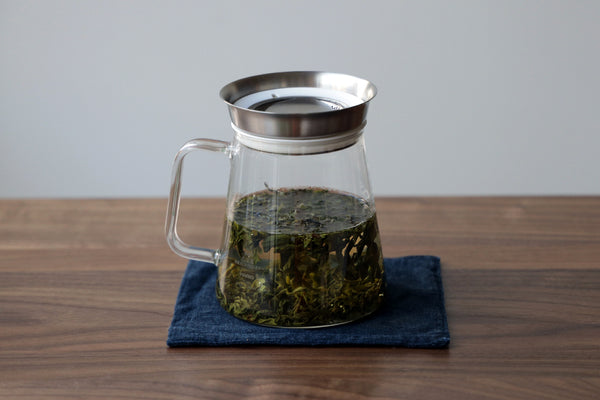 A glass tea brewing pitcher with loose tea leaves in water.