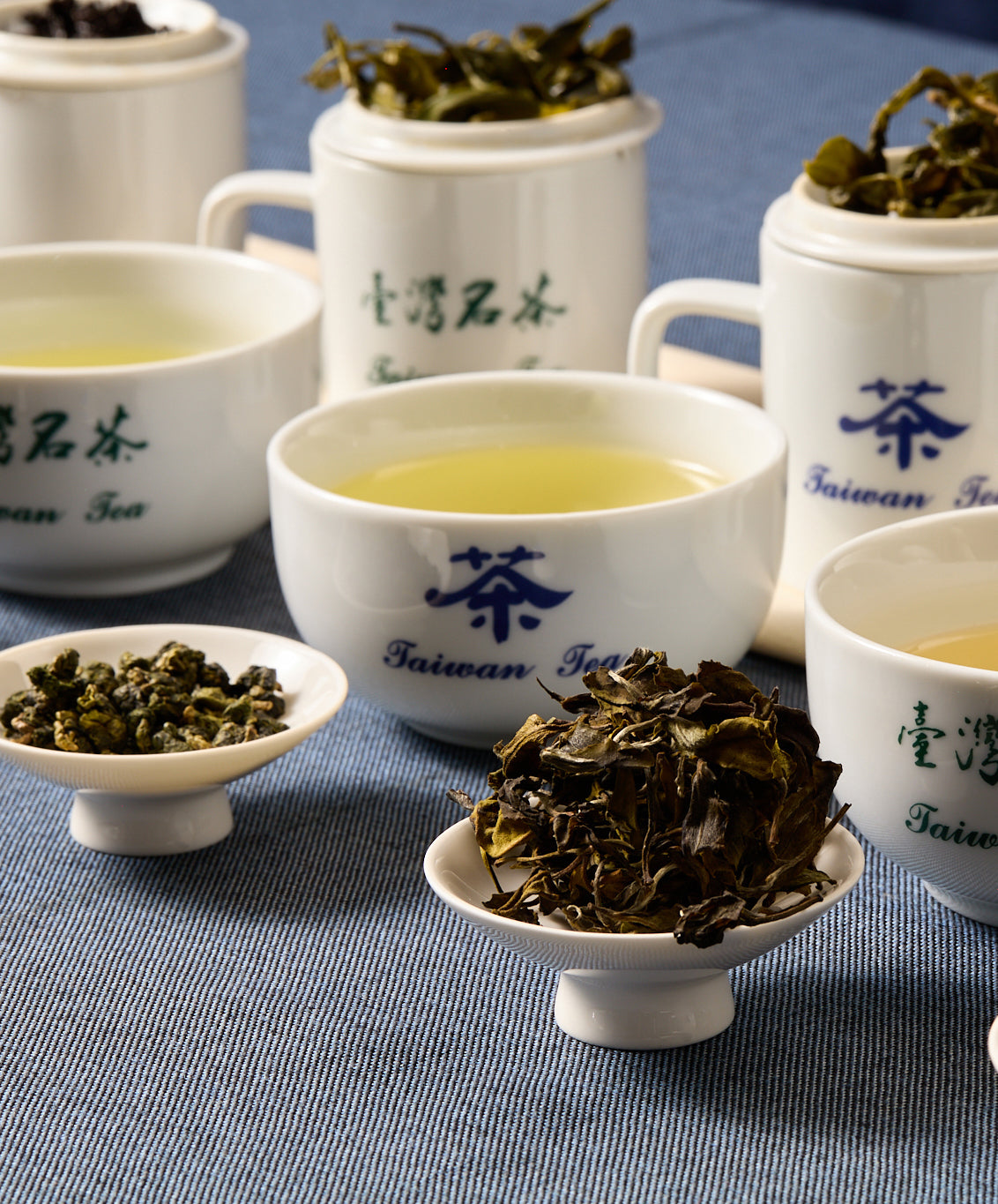 A tea-tasting setup with white ceramic cups labeled "Taiwan Tea," filled with tea and surrounded by loose tea leaves on small dishes.