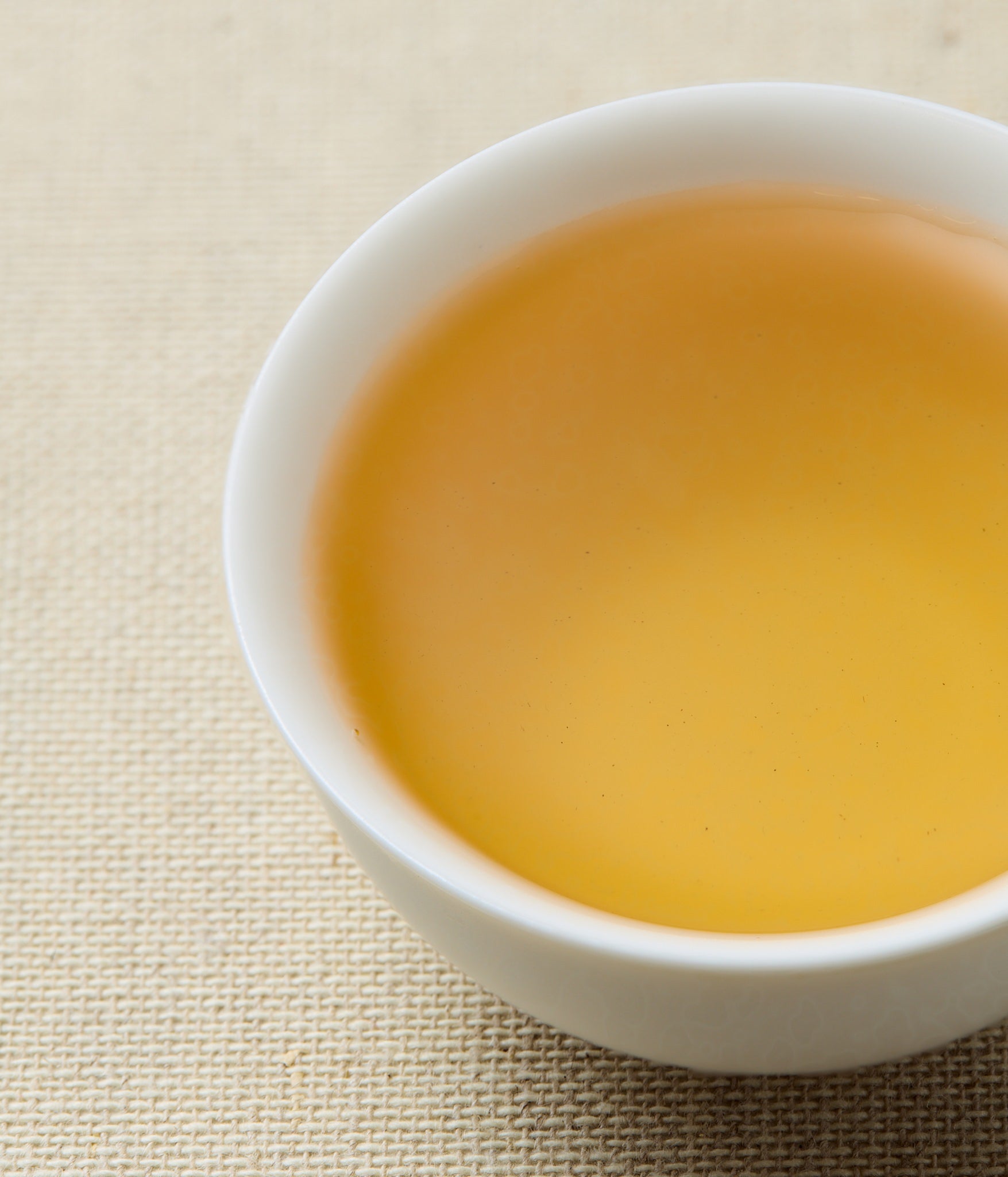 A close-up of a white ceramic cup filled with light golden tea, set on a beige woven fabric surface.