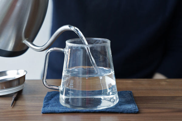 A kettle pouring hot water into a tea brewing pitcher with loose leaves.