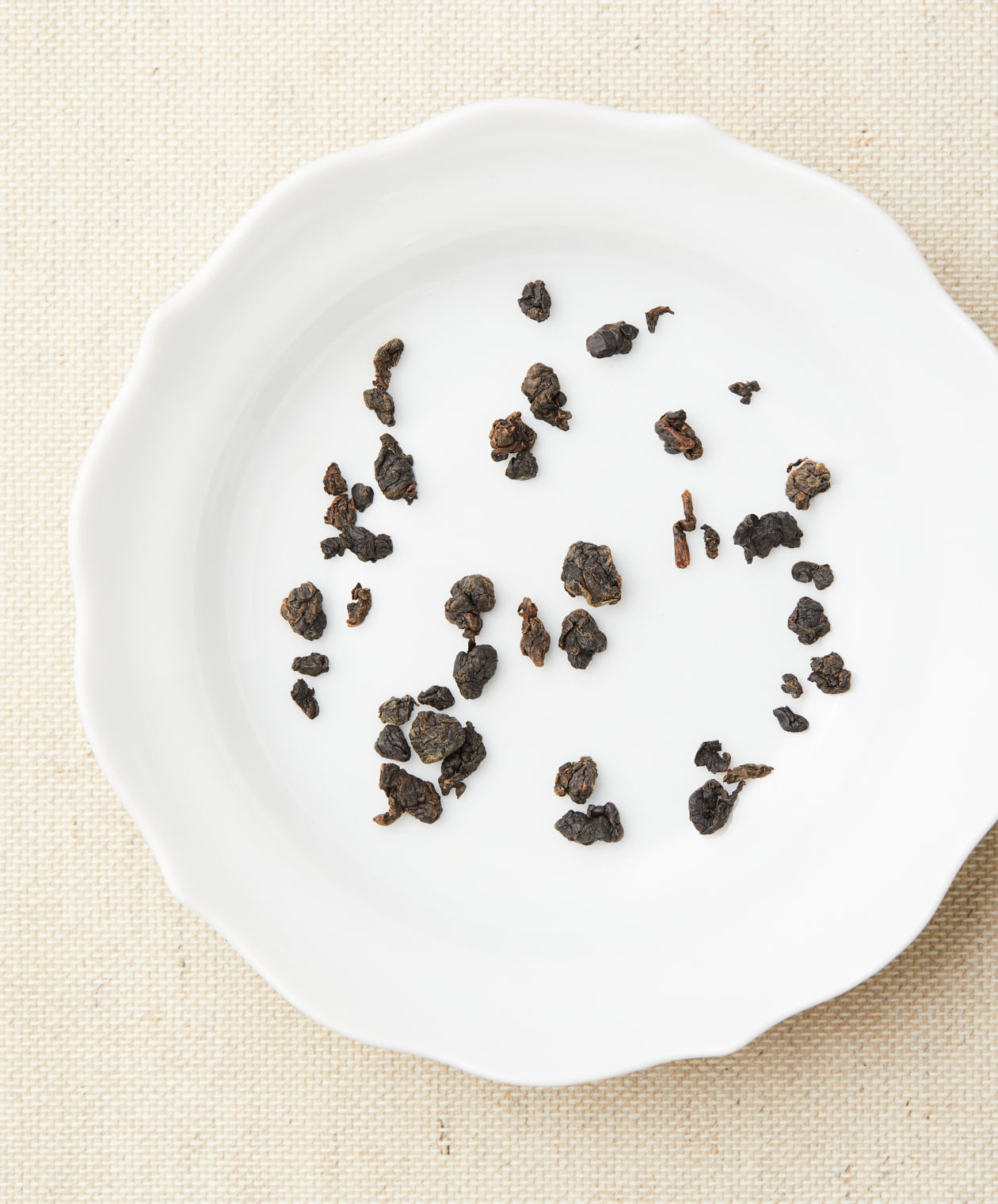 Loose-leaf tea in curled, dark pieces arranged on a white plate against a beige fabric background.