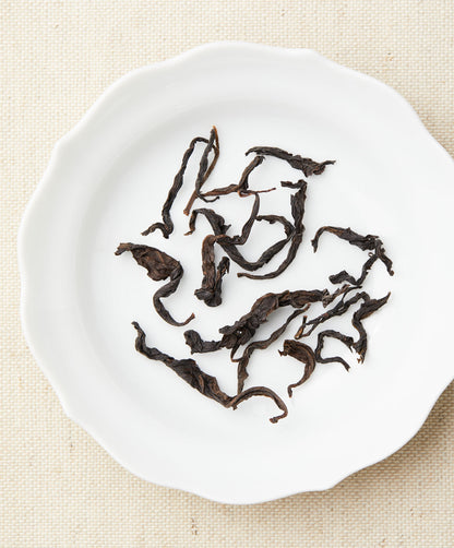 Twisted Loose-leaf tea in curled, dark pieces arranged on a white plate against a beige fabric background.