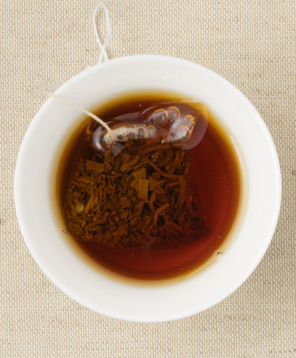 A cup of steeped brownish tea with a pyramid-shaped tea sachet containing loose leaves, set against a beige fabric background.