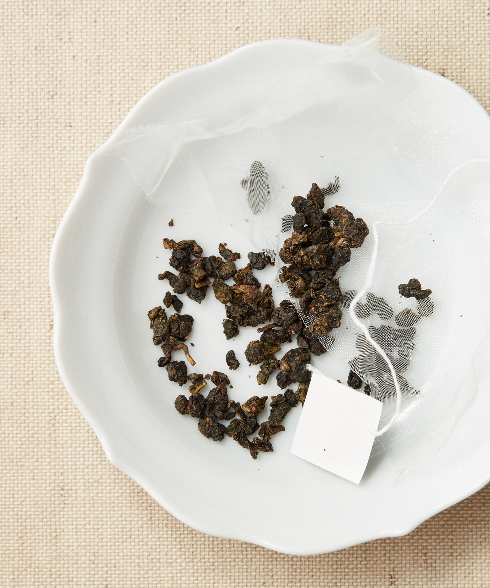 A pyramid-shaped tea sachet with loose leaves scattered around it on a white plate, set against a beige fabric background.
