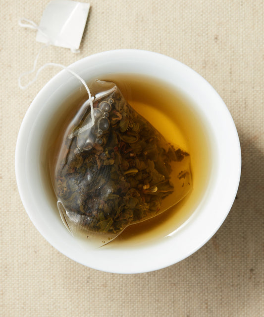A cup of steeped tea with a pyramid-shaped tea sachet containing loose leaves, submerged in the liquid.
