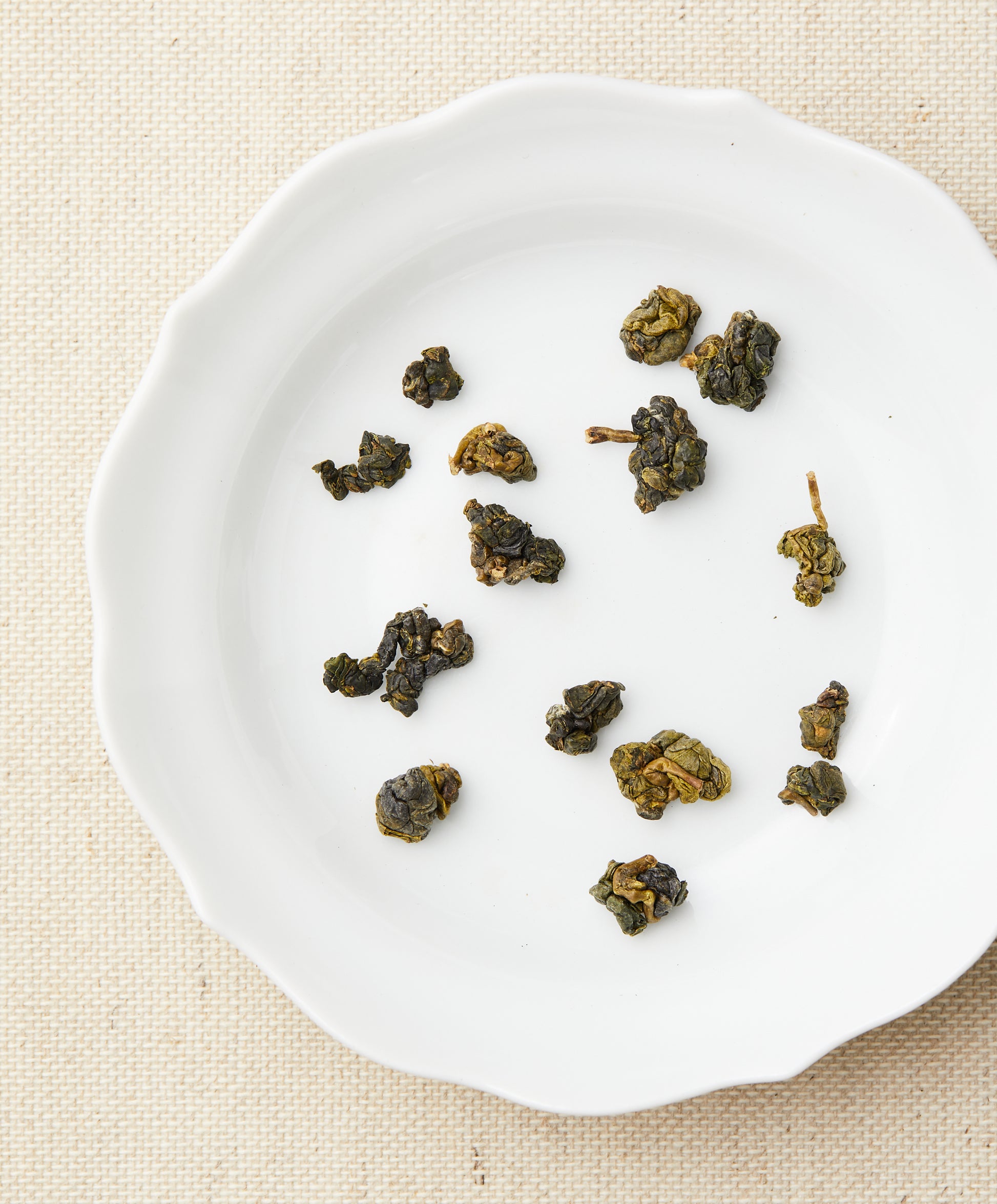Loose-leaf tea pieces arranged on a white plate, showing tightly rolled green tea leaves against a beige fabric background.