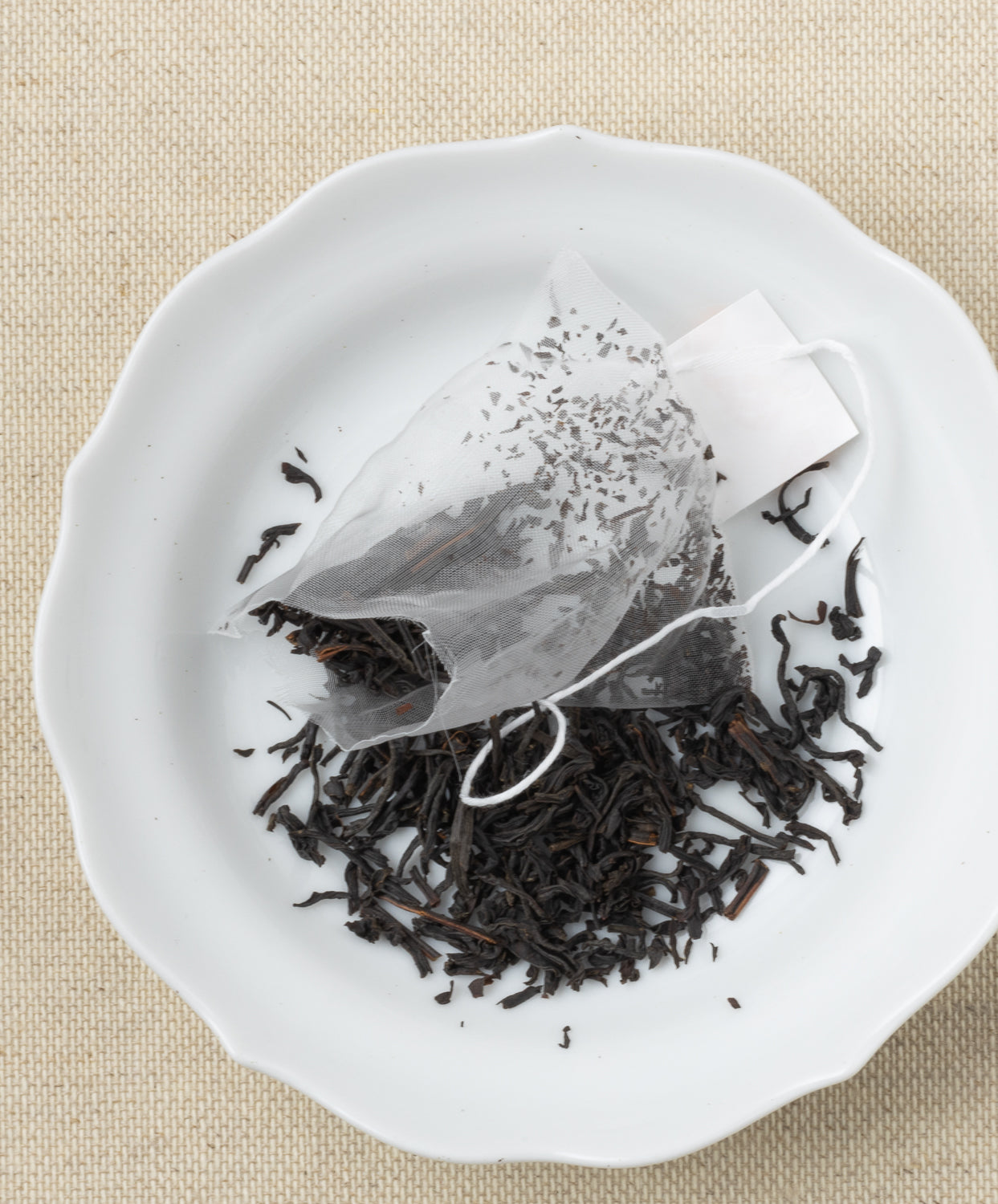 A pyramid-shaped tea sachet with loose leaves scattered around it on a white plate, set against a beige fabric background.