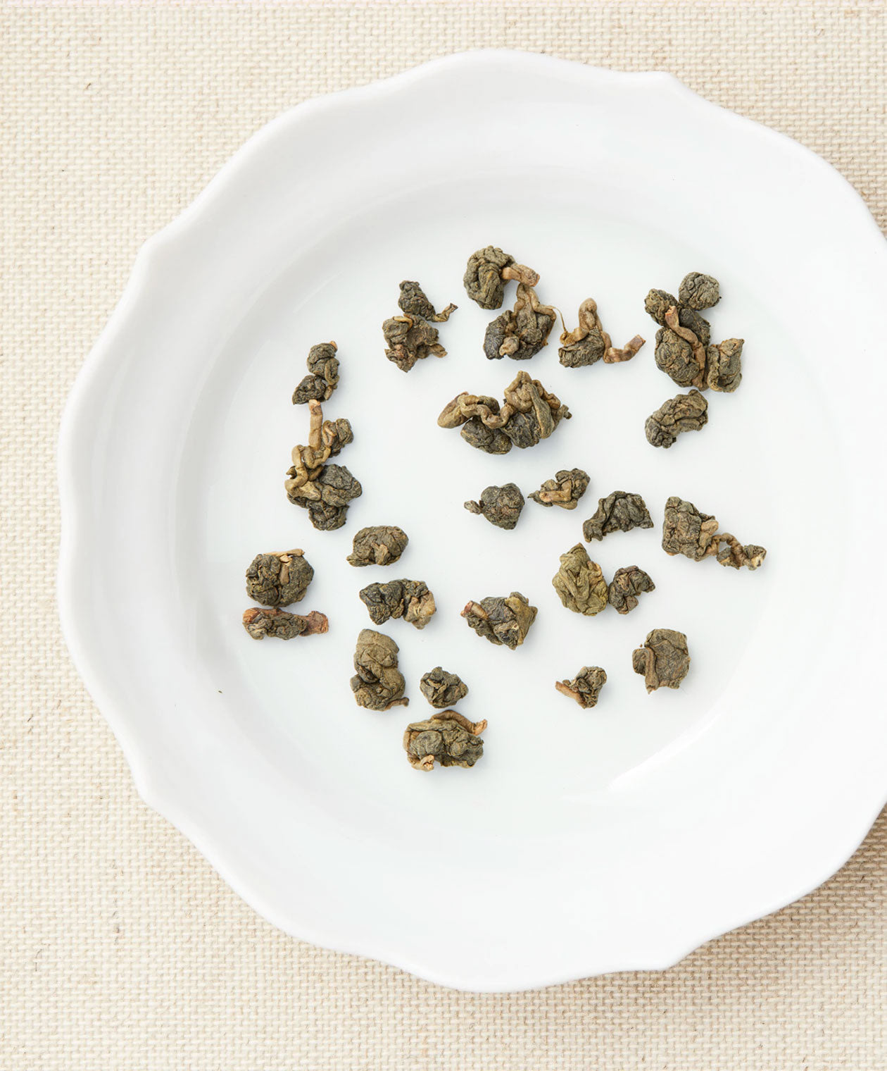 Loose-leaf tea pieces arranged on a white plate, showing tightly rolled green tea leaves against a beige fabric background.