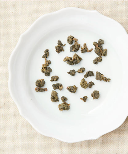 Loose-leaf tea pieces arranged on a white plate, showing tightly rolled green tea leaves against a beige fabric background.