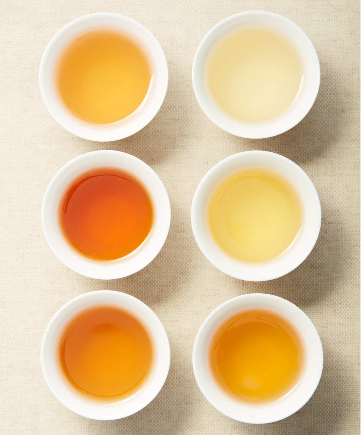 Six white teacups filled with tea of varying shades, ranging from pale yellow to deep amber, displayed on a beige textured background.