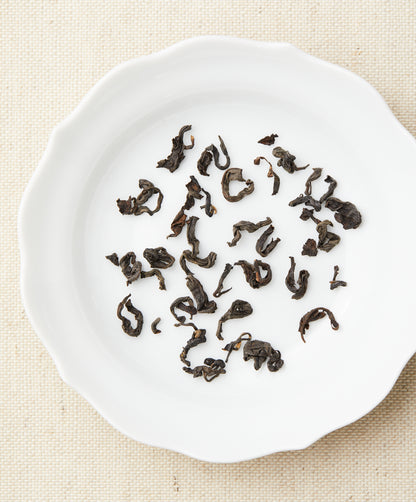 Loose-leaf tea in curled, dark pieces arranged on a white plate against a beige fabric background.