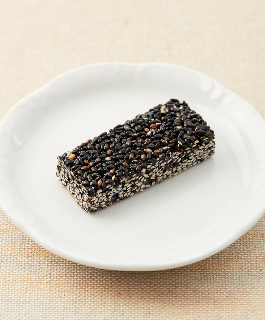 A side view of a black sesame seed bar on a white plate against a beige fabric background.