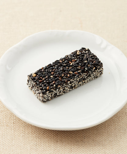 A side view of a black sesame seed bar on a white plate against a beige fabric background.